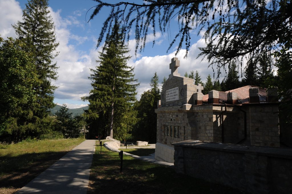 Stone monument with a cross on top, along a paved walkway bordered by tall fir trees