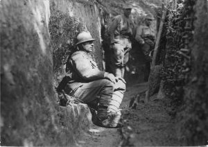 Soldier with helmet sitting in an earthen trench, with two other servicemen in the background