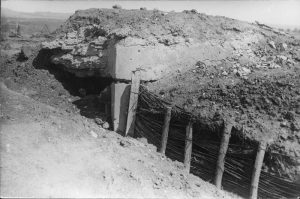 Military trench fortified with wooden posts and woven twigs, next to a concrete structure covered in earth