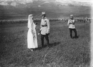 A woman in white clothing with a veil talking to a military officer on an open field in front of troops
