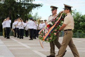 Two soldiers carrying a large commemorative flower wreath, in front of a military brass band
