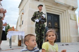 Two children seated outside Mărăști Mausoleum, near a guard soldier in camouflage uniform